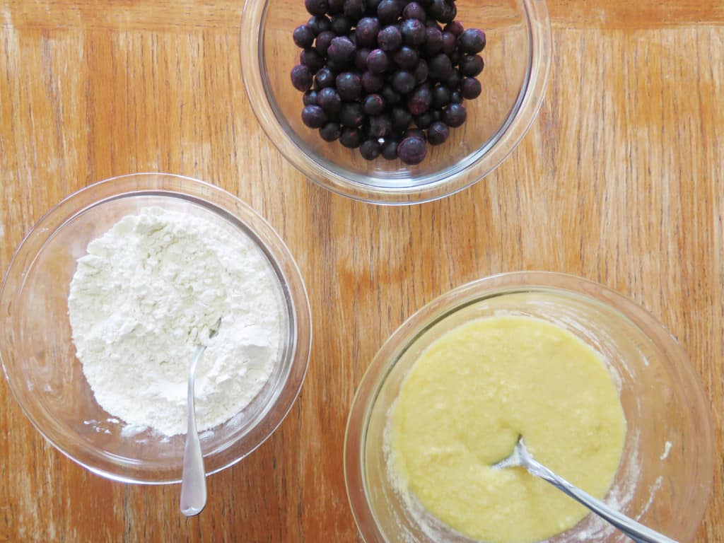 Flour mixture, egg mixture, and frozen blueberries on a table.