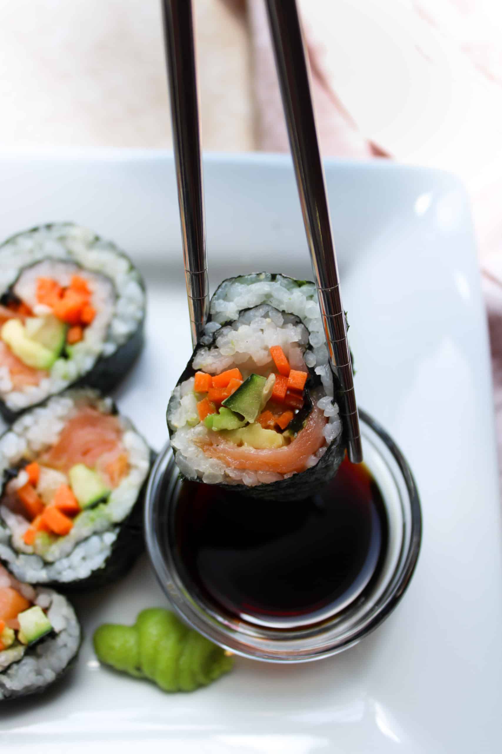 A sushi roll being held up with chop sticks over a dipping bowl filled with soy sauce.