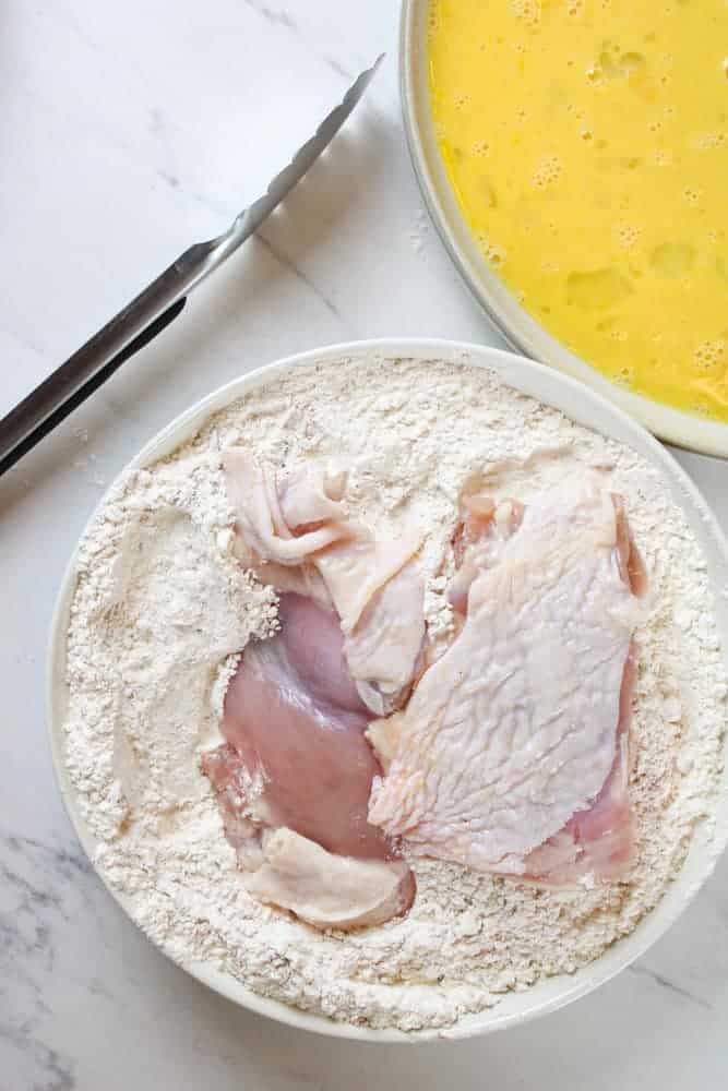 Raw chicken in a bowl of flour with a pair of tongs and a bowl of whisked eggs near by.