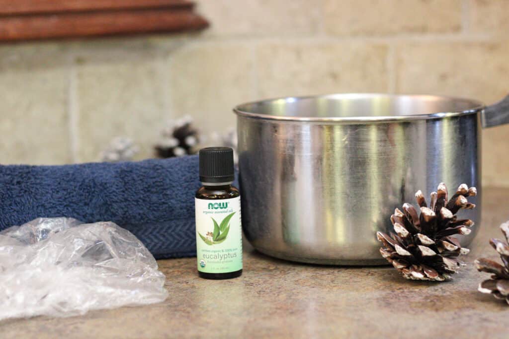 Side image of eucalyptus oil, pot, towel, and shower cap on a table with a pinecone. 