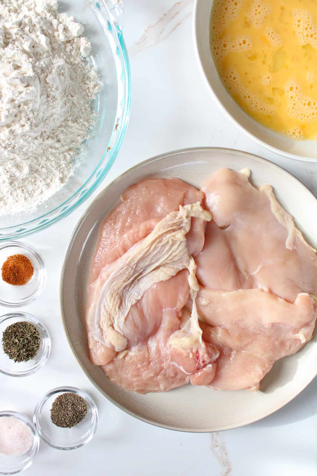 Chicken breast on a plate surrounded by seasonings, flour, and whisked eggs. 