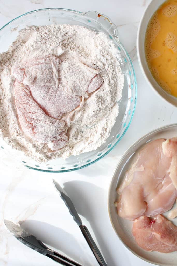 Chicken breast coated in flour in a pie dish, surrounded by whisked eggs, and raw chicken breast on a plate.