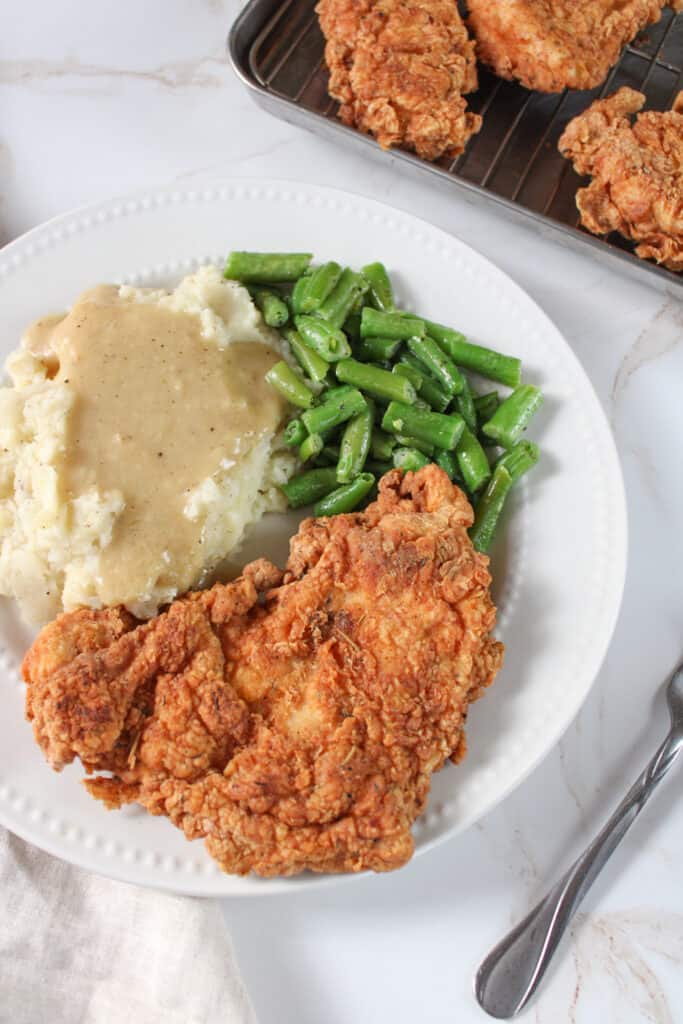 Side view of chicken fried chicken on a plate with mashed potatoes gravy, and green beans.