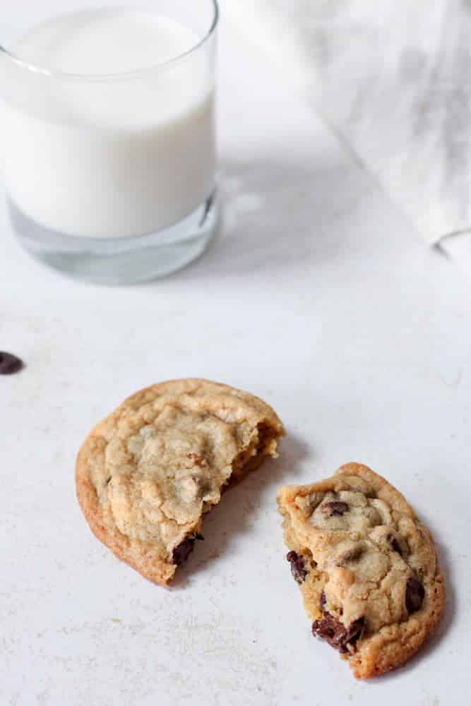 One broken chocolate chip cookie on a table with a glass of milk.
