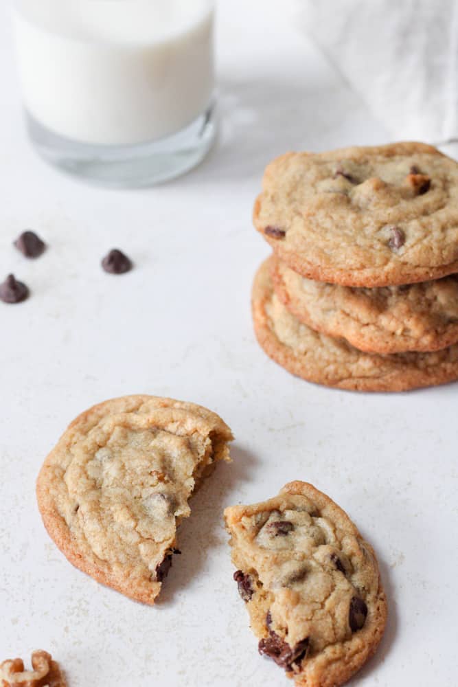 3/4 view of a broken chocolate chip cookie with a glass of milk and a stack of cookies in the background.