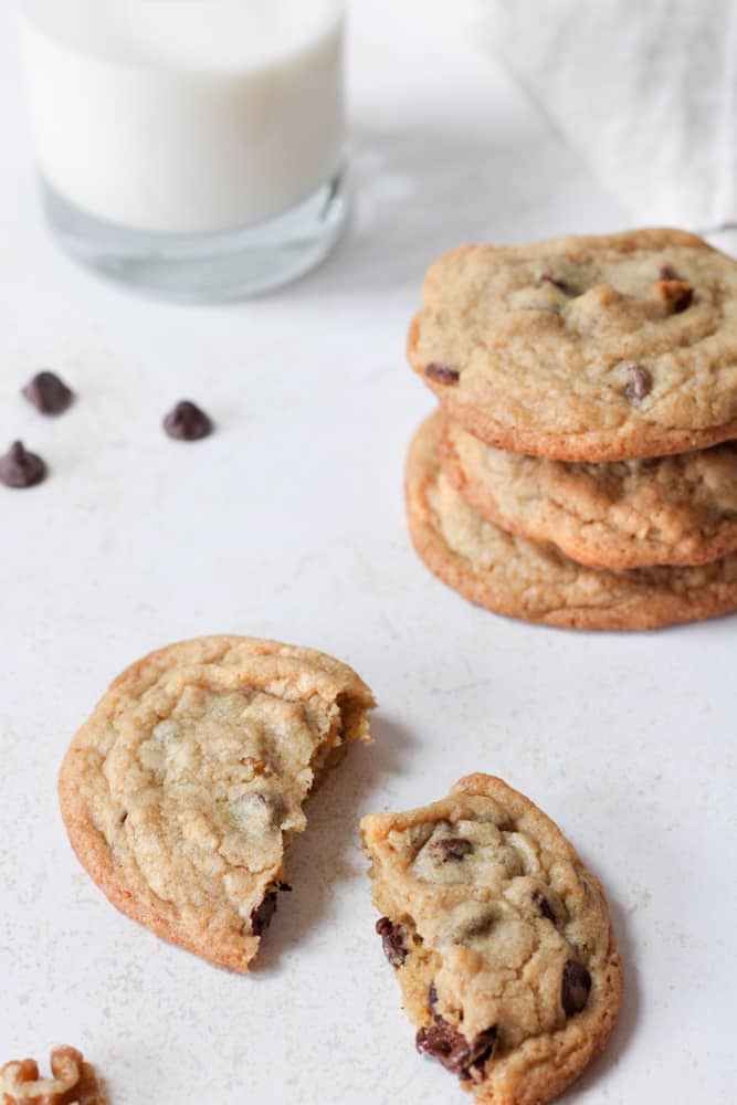 ¾ view of a broken chocolate chip cookie with a glass of milk and a stack of cookies in the background.