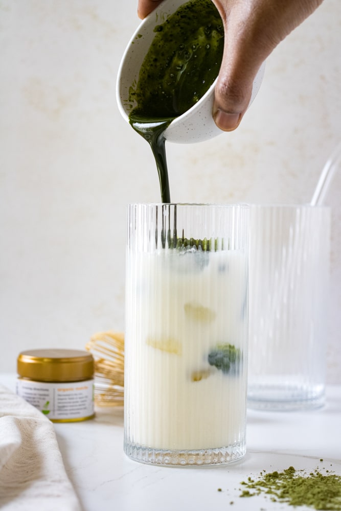Woman's hand pouring matcha mixture into glass with milk and ice.