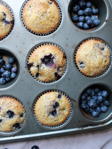 Blueberry muffins in a muffin pan with fresh blueberries.