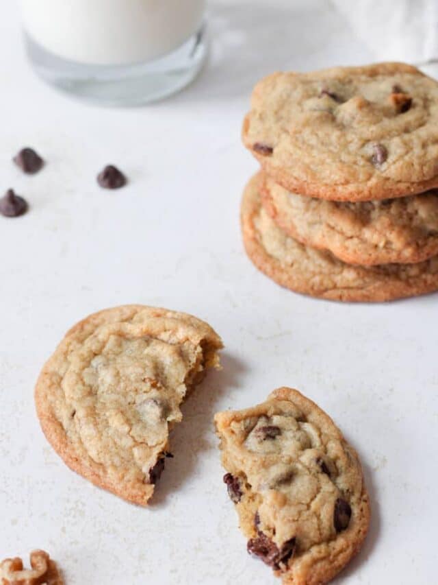 3/4 view of a broken chocolate chip cookie with a glass of milk and a stack of cookies in the background.