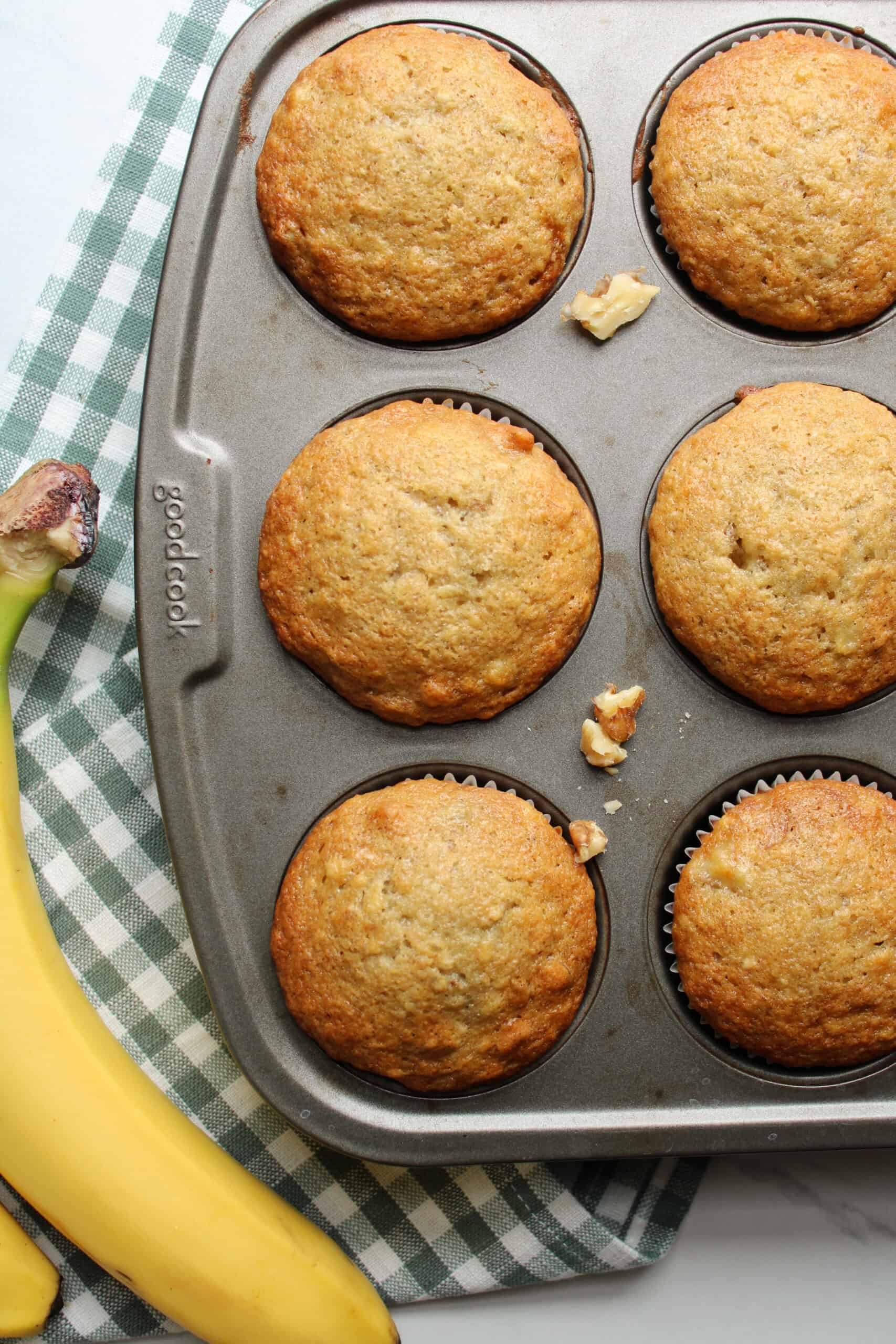 Moist banana nut muffins in a muffin pan with nuts and a banana on a counter.