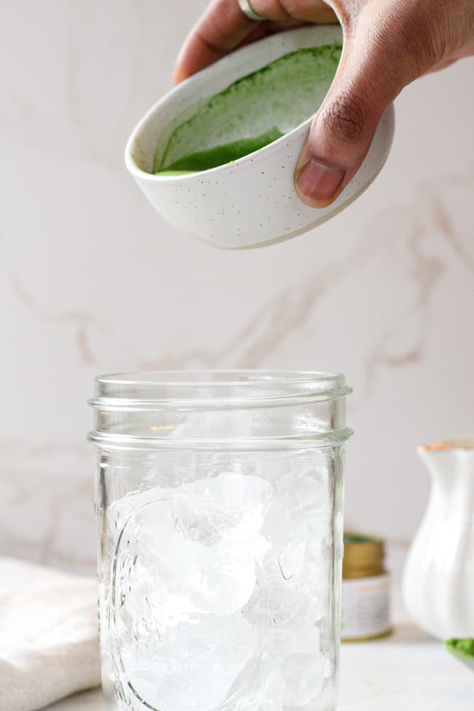 Side view of a mason jar filled with ice. Woman's hand holding matcha bowl over jar.