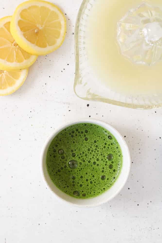 Overhead view of matcha powder in a bowl with lemons and lemon juice on a table.