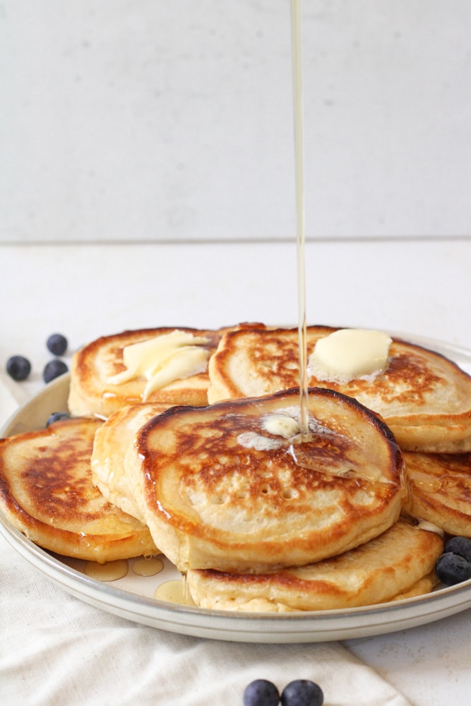 Pouring image of maple syrup over a stack of pancakes with fresh blueberries on a plate.