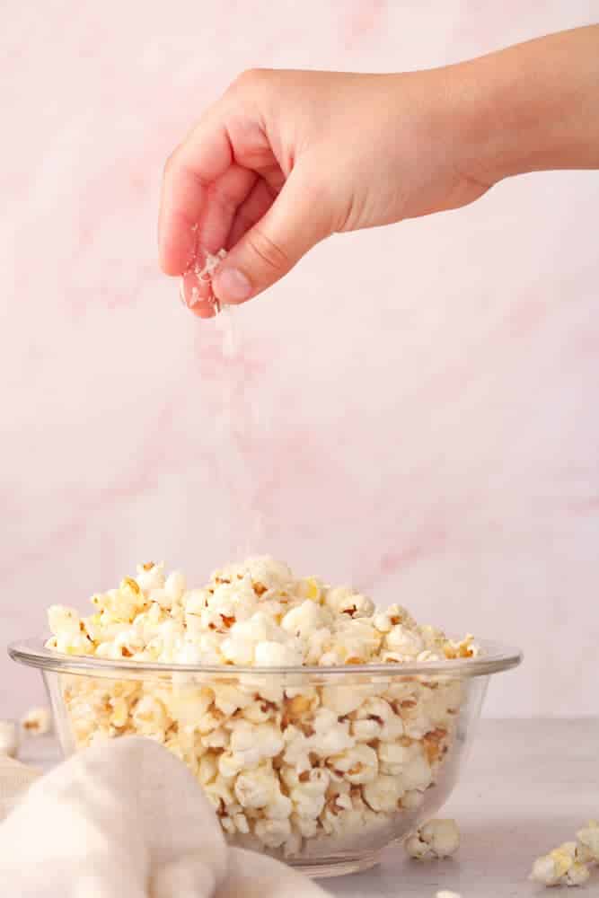 Boy's hand sprinkling parmesan cheese on bowl of popped popcorn. 