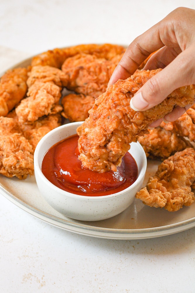 Overhead image of woman's hand dipping chicken tender into ketchup.