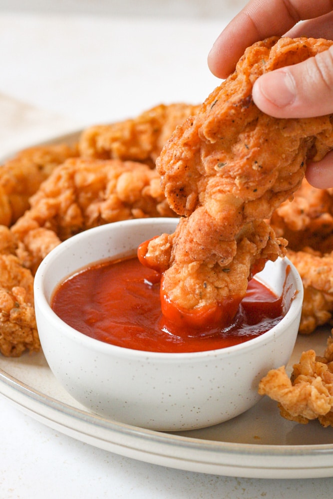 Boy's hand dipping chicken tender into bowl of ketchup.