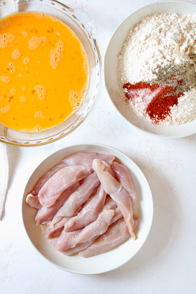 Sliced chicken strips on a plate with seasoned flour mixture and egg mixture near by.