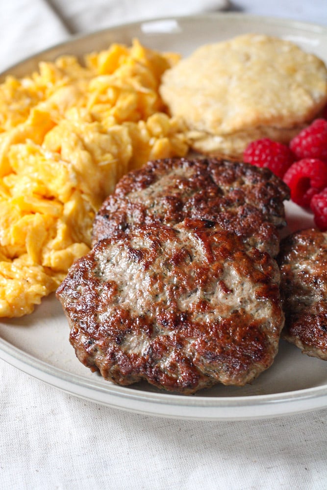 Close up image of breakfast sausage patties on a plate with fruit, eggs, and biscuits.