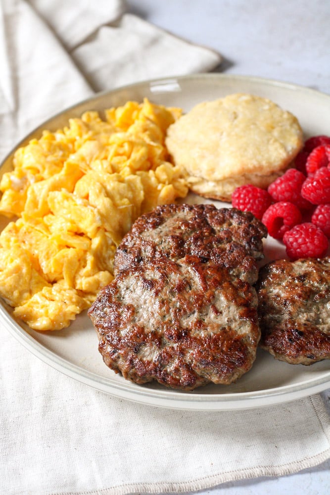Close up a plate of lamb sausage patties with eggs, fruit, and biscuits.