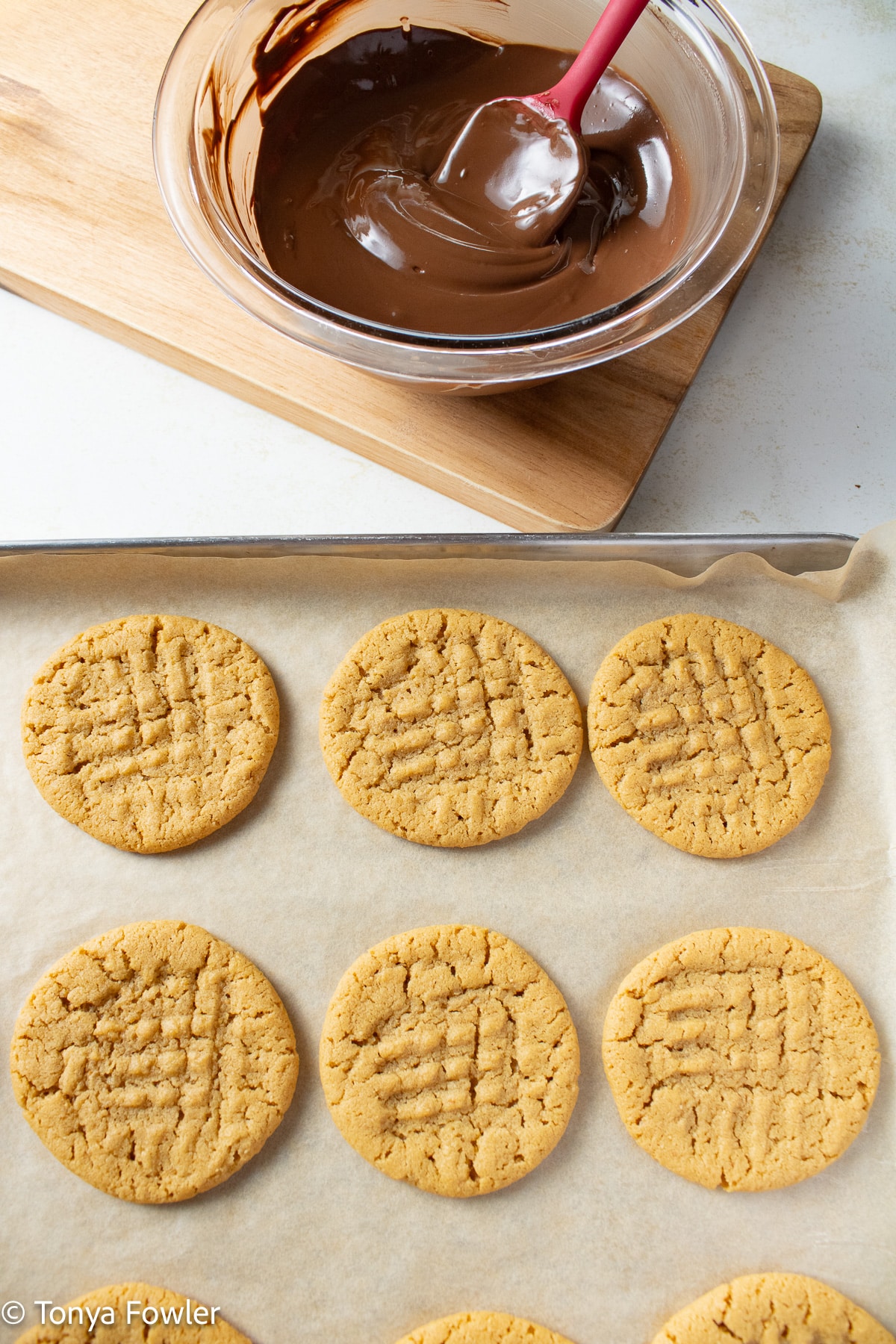 Baked cookies on a sheet pan with melted chocolate in a bowl.