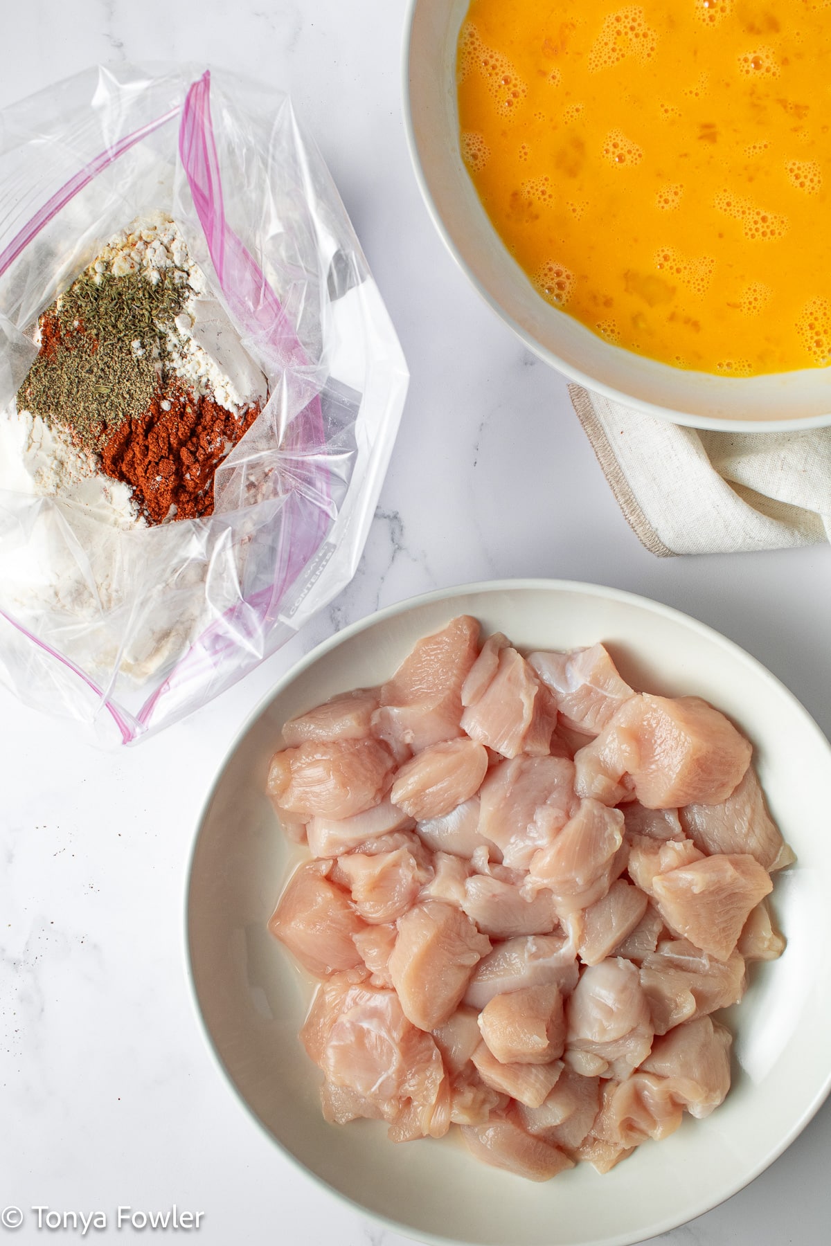 Cut chicken in a plate, seasonings in a plastic bag and eggs whisked in a shallow bowl nearby.