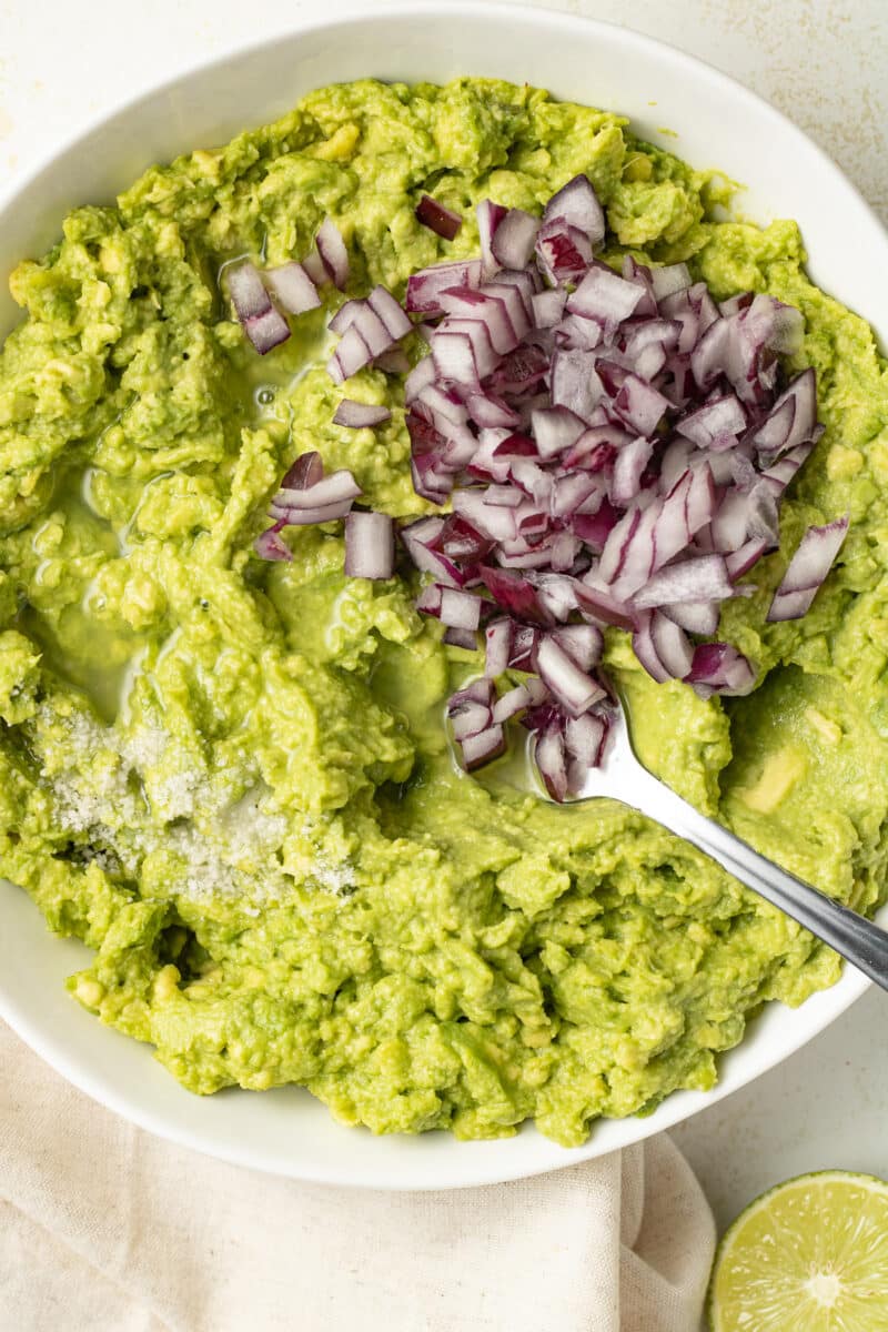 Mashed avocado in a bowl topped with alt and diced red onions.