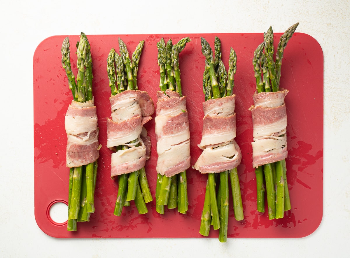 Raw bacon wrapped asparagus on a cutting mat.