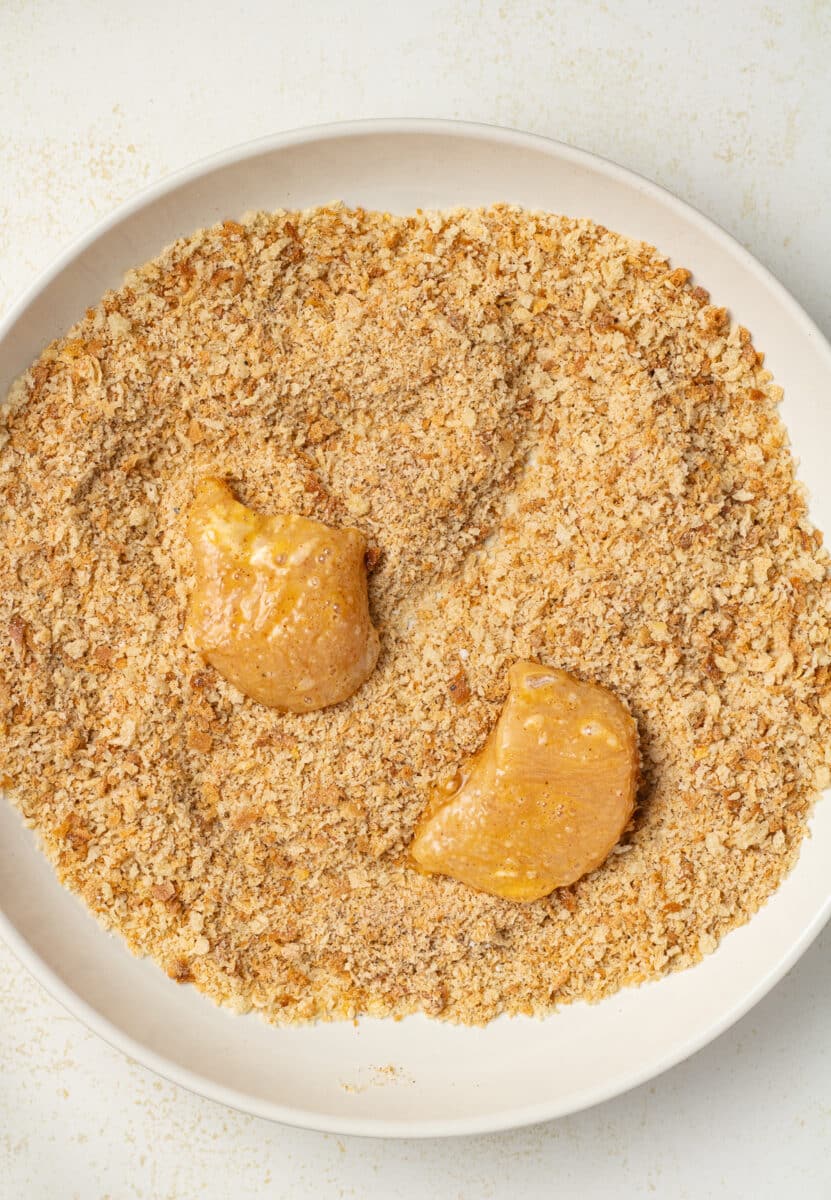Chicken piece coated in egg in a bowl of bread crumbs.
