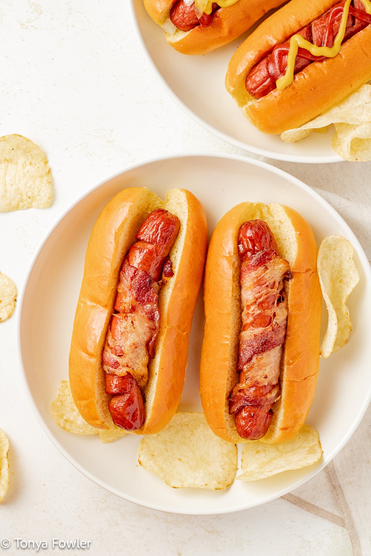 Bacon hot dogs on buns on a plate with chips.