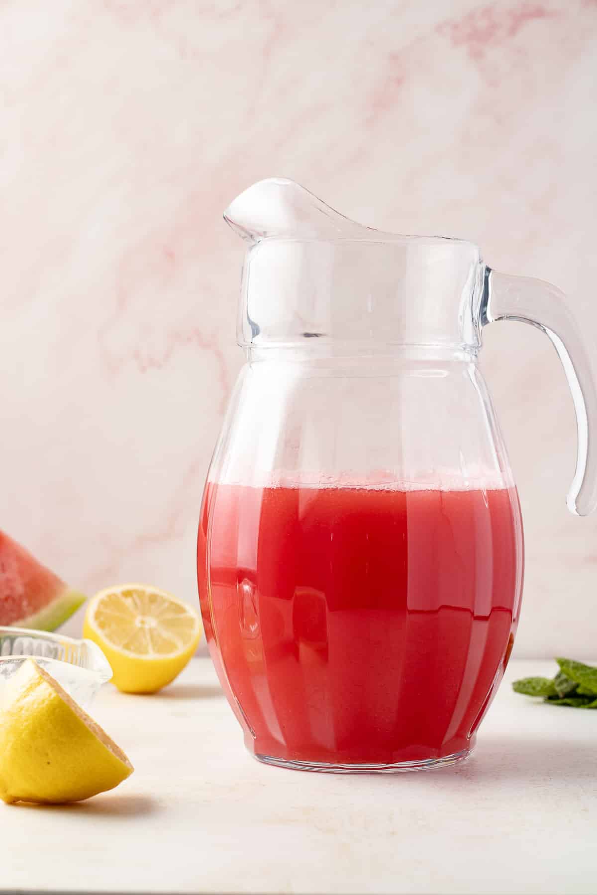Watermelon mint lemonade in a pitcher with lemon halves on a counter.