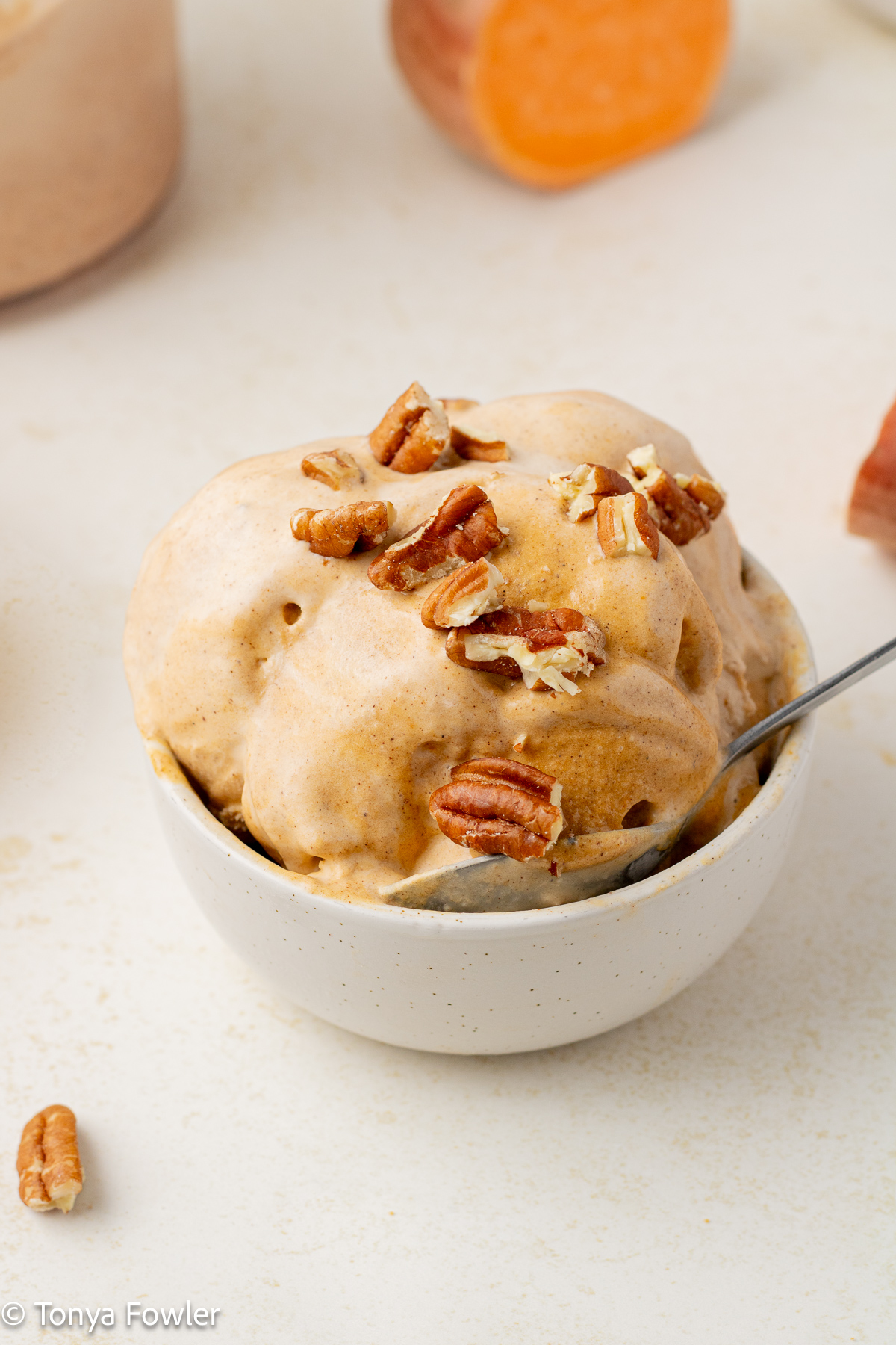 3/4 view of sweet potato ice cream in bowl with a spoon topped with nuts surrounded by nuts and a half of a sweet potato.