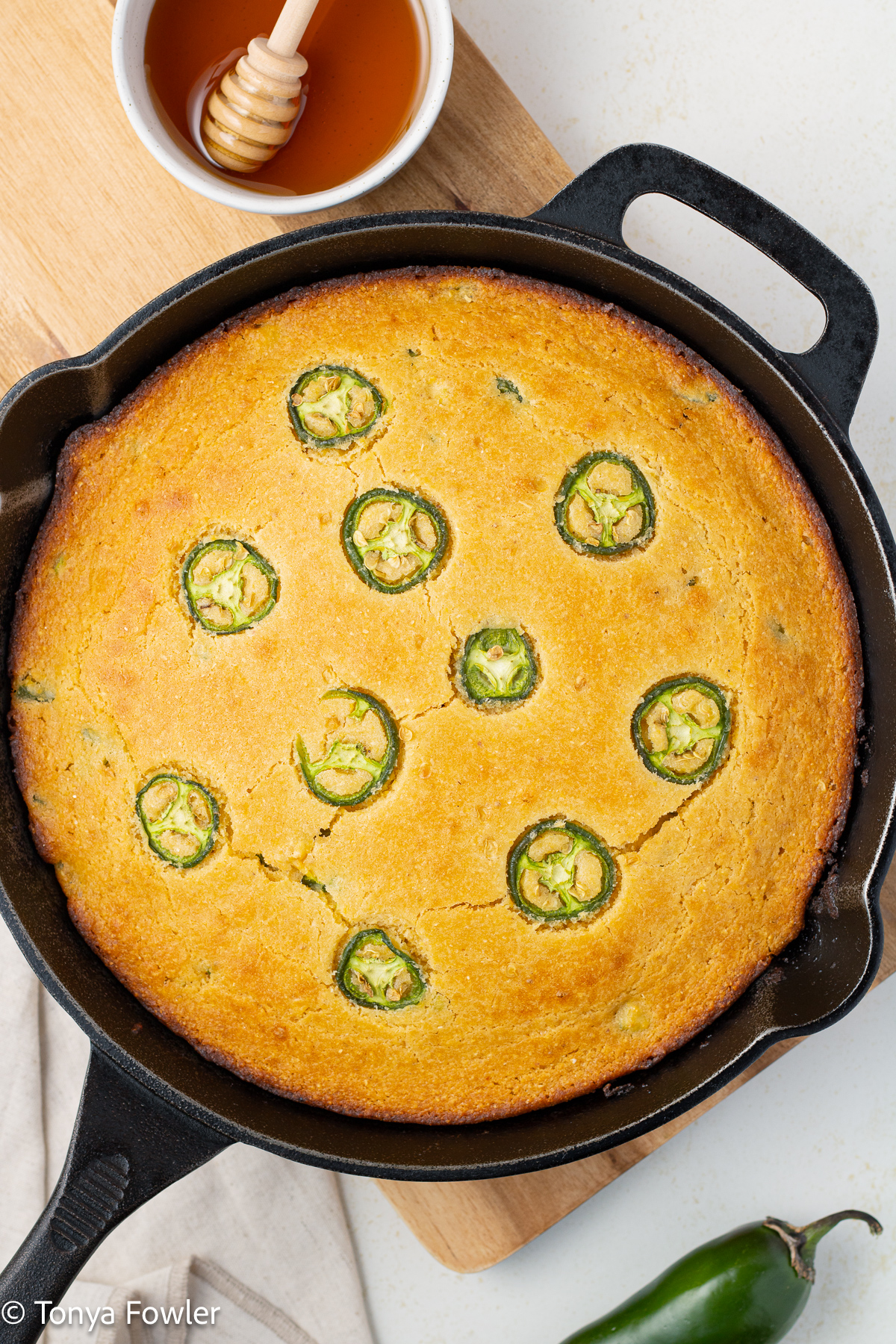Overhead view of jalapeno honey cornbread in a skillet surrounded by honey and jalapenos.