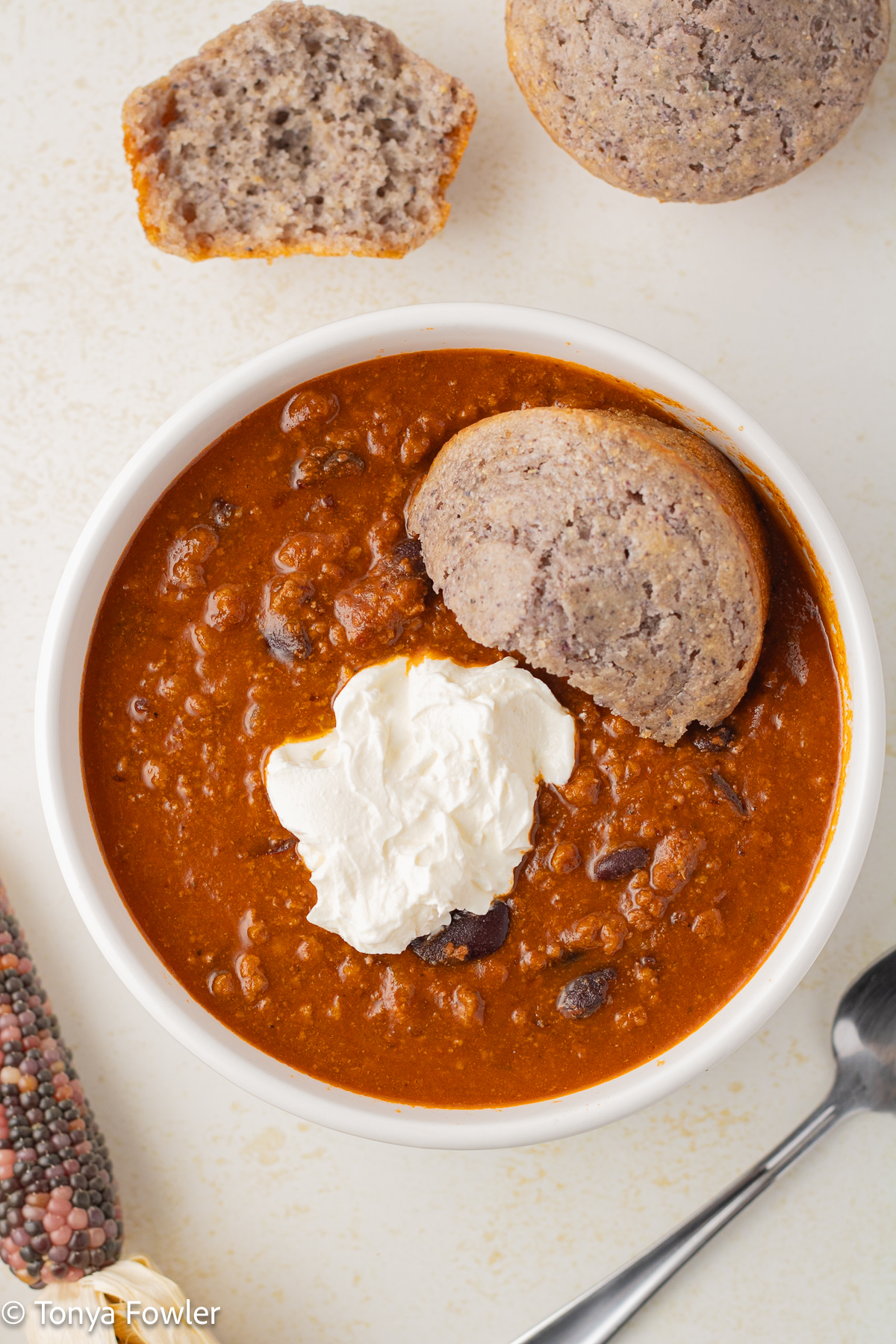 Overhead view of chili in a bowl topped with sour cream and a cornmeal muffin.