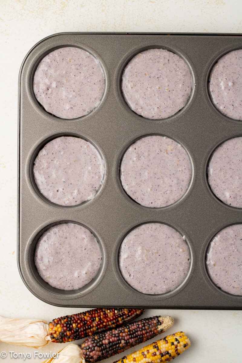 Cornmeal batter in a muffin tin with decorative corn cobs on the table.