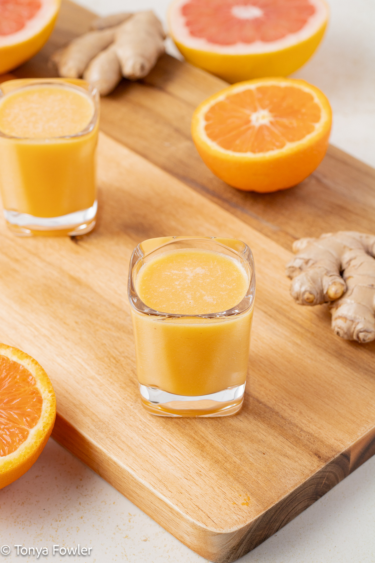 3/4 view of Citrus Grapefruit Ginger Wellness Shots on a cutting board surrounded by ginger, and fresh oranges and grapefruit.