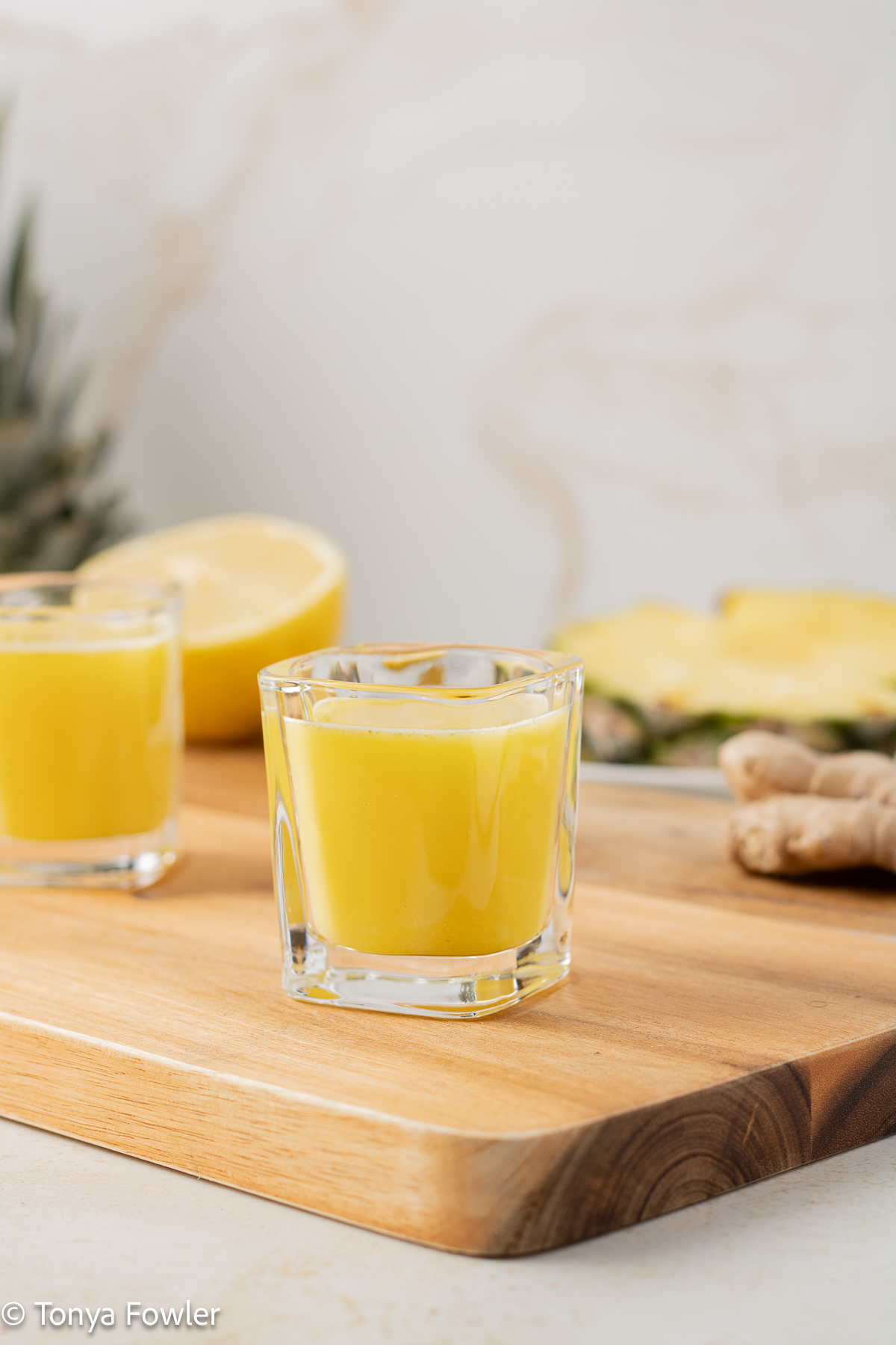 Side view of Pineapple Ginger Shots in glasses with a pineapple, sliced lemon and ginger root near by on a cutting board.
