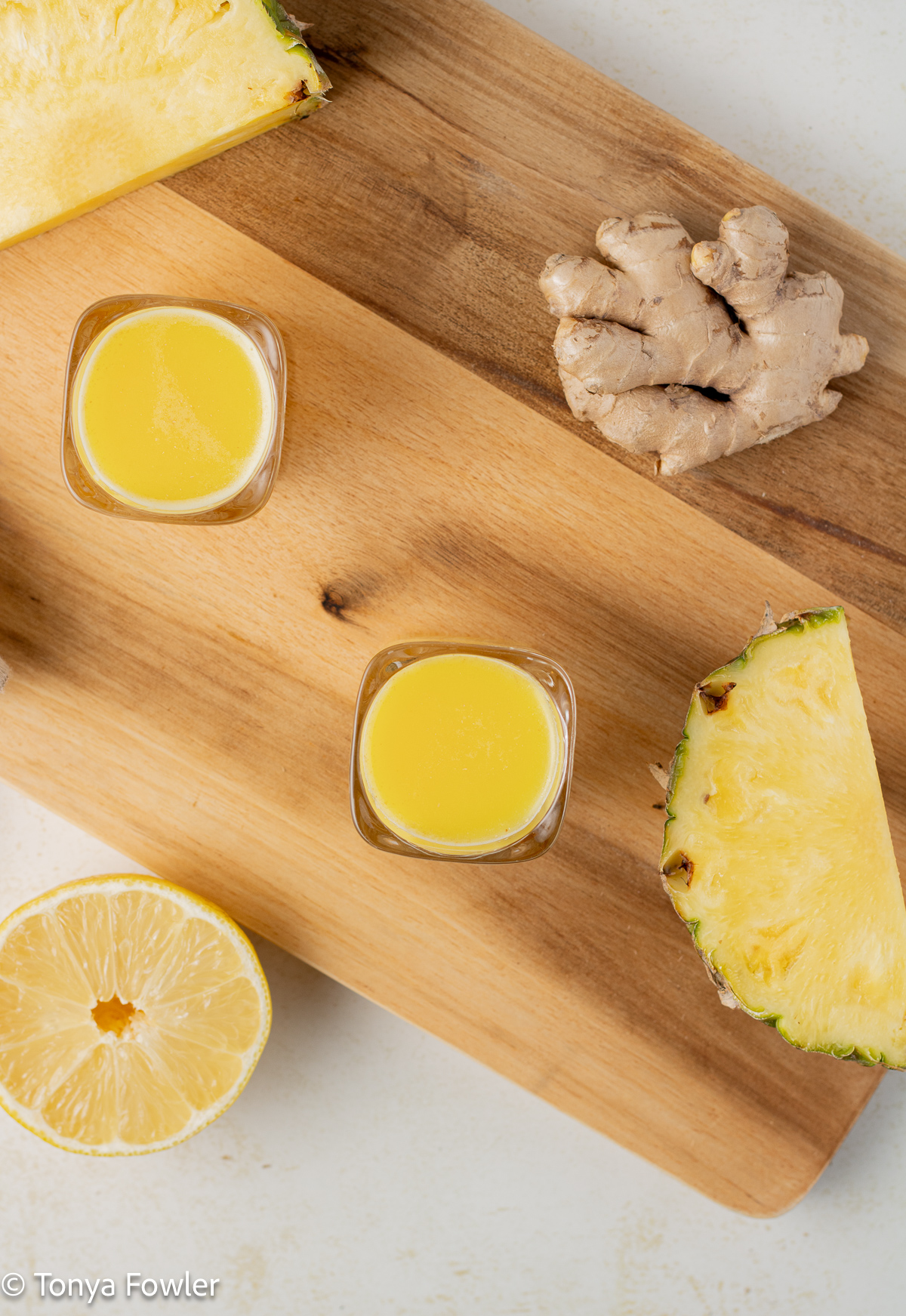Overhead view of ginger shots on a cutting board with pineapple slices, ginger root and a half of lemon.