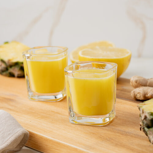 Side view of pineapple ginger shots sitting on a cutting board with slices of pineapple and lemon.
