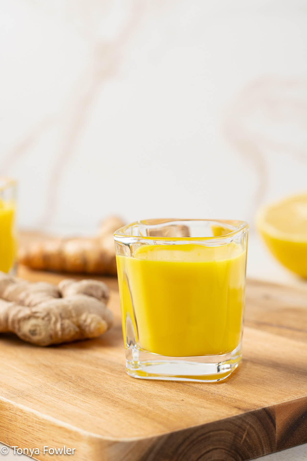 Side view of a Turmeric Ginger Shot on a cutting board with ginger and turmeric roots in the background.