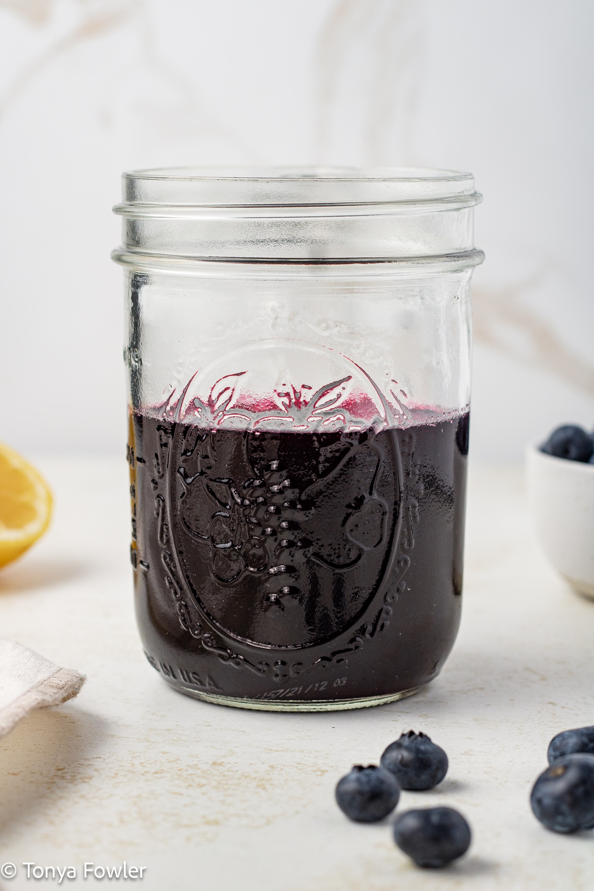 Side view of blueberry syrup in a mason jar with fresh blueberries on a table.