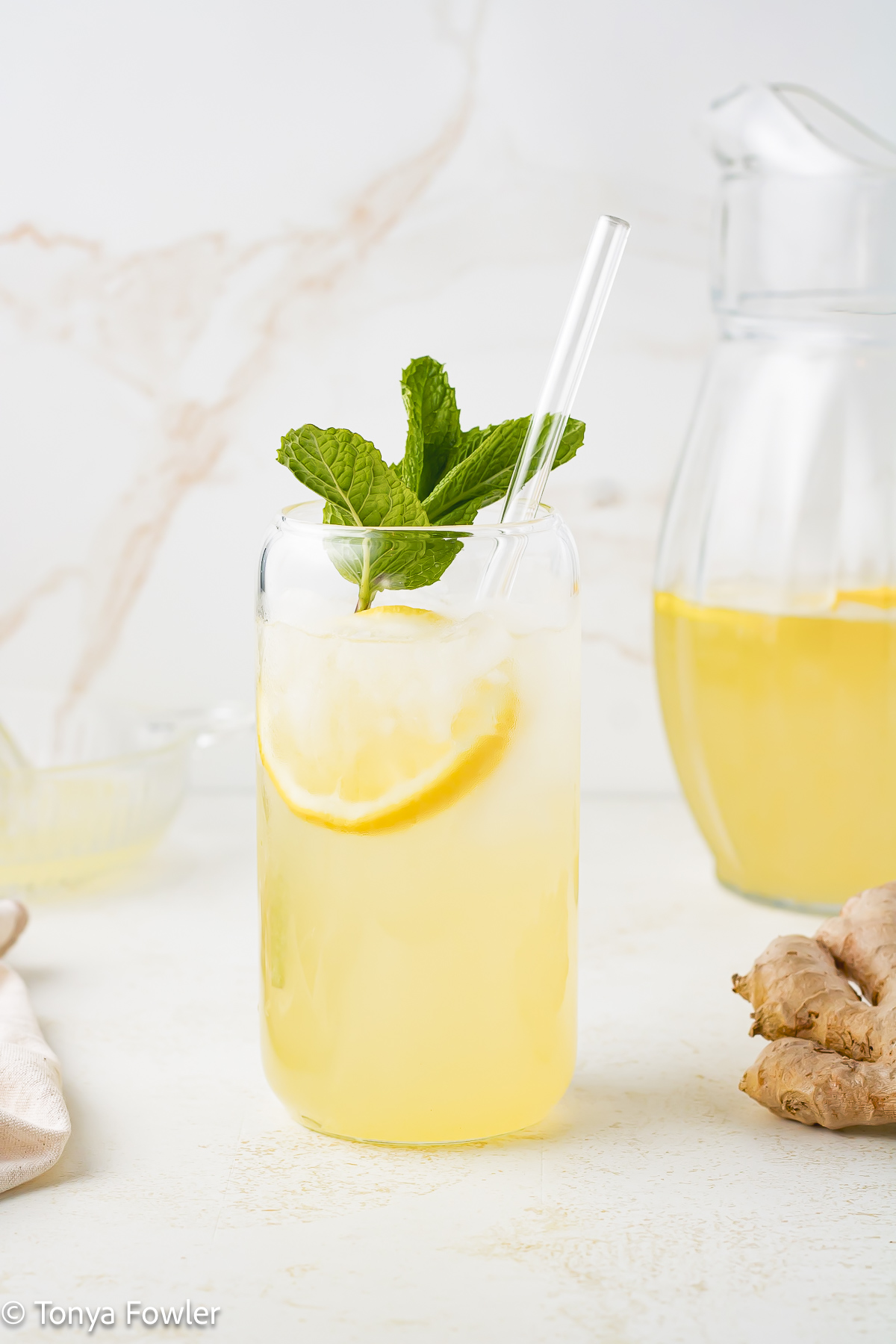 Straight on view of a glass of lemonade with fresh mint and ginger root near by.