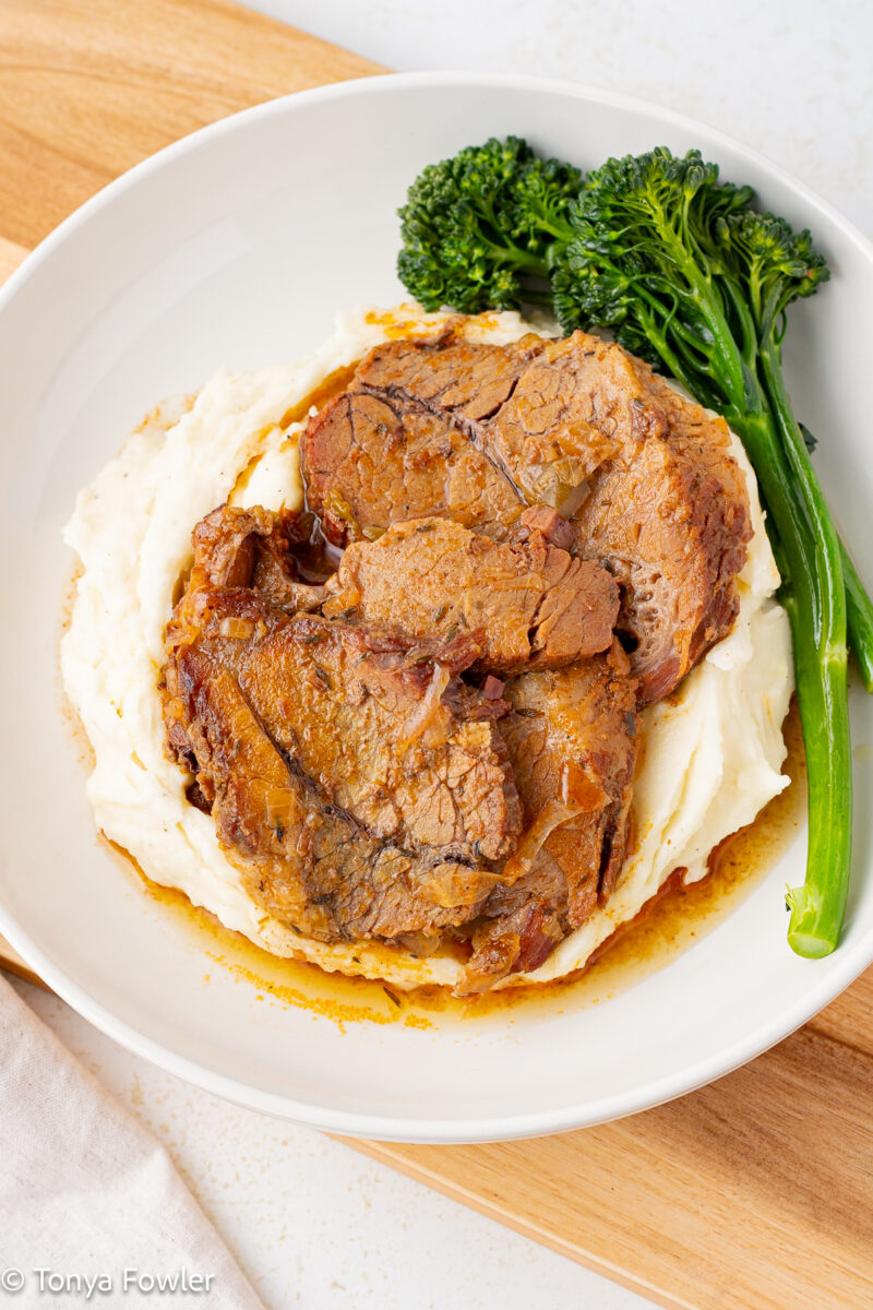 3/4 view of sliced brisket on a bed of mashed potatoes with a side of baby broccoli.