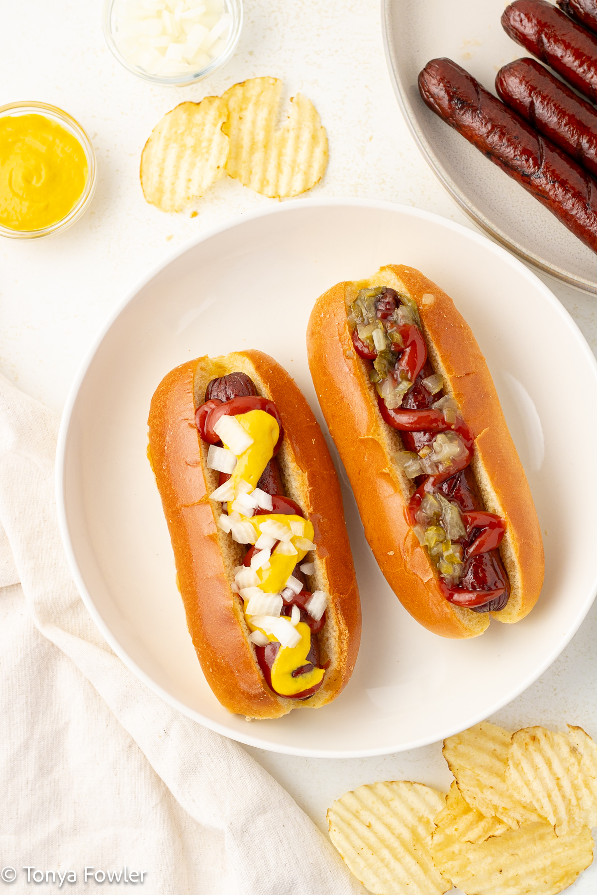 Overhead image of smoked hot dogs topped with toppings in buns with potato chips near by.