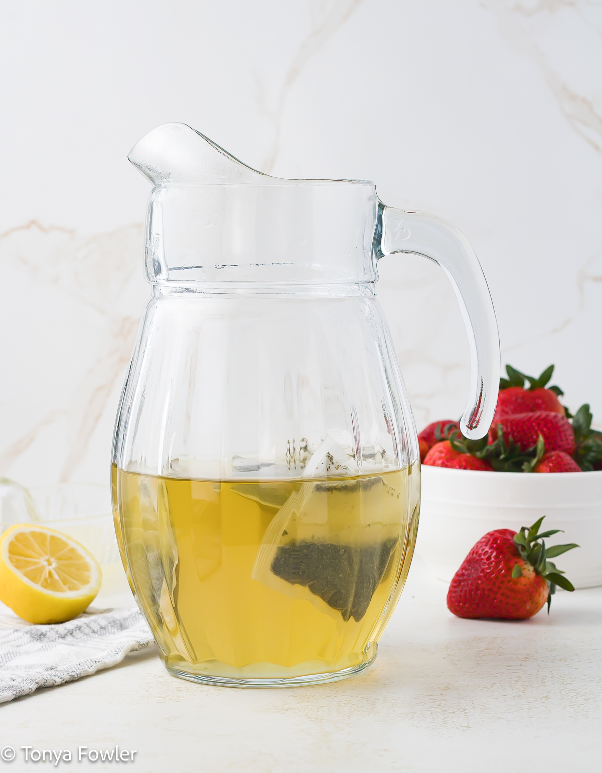 Tea steeping in a pitcher with fresh strawberries and lemons in the background.