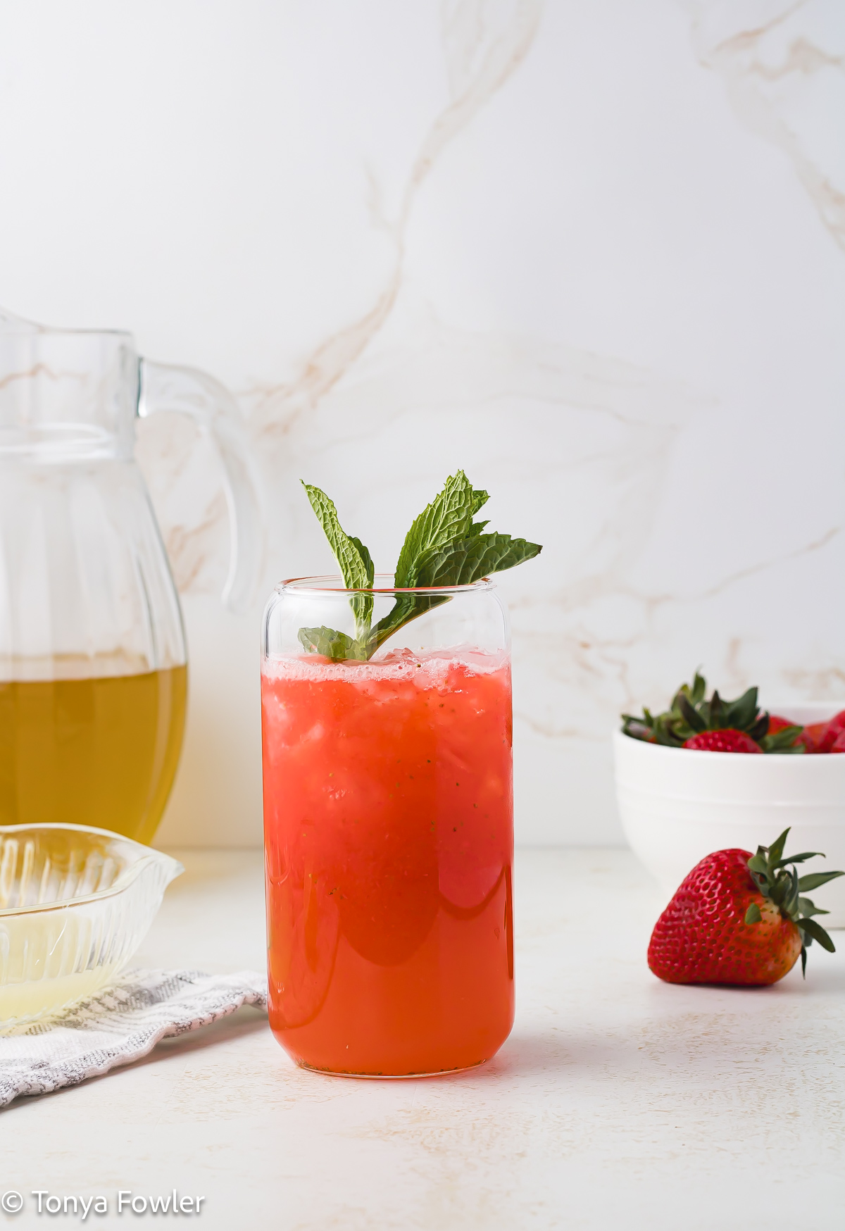 Straight on view of strawberry lemonade in a glass with a pitcher of tea in the background.