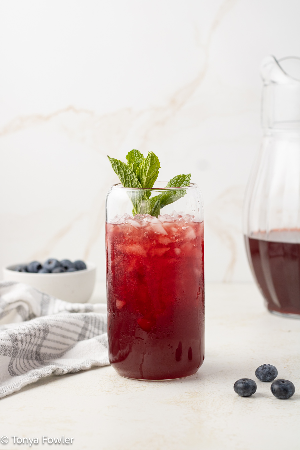 Straight on image of blueberry iced tea in a glass with mint and blueberries on the table.
