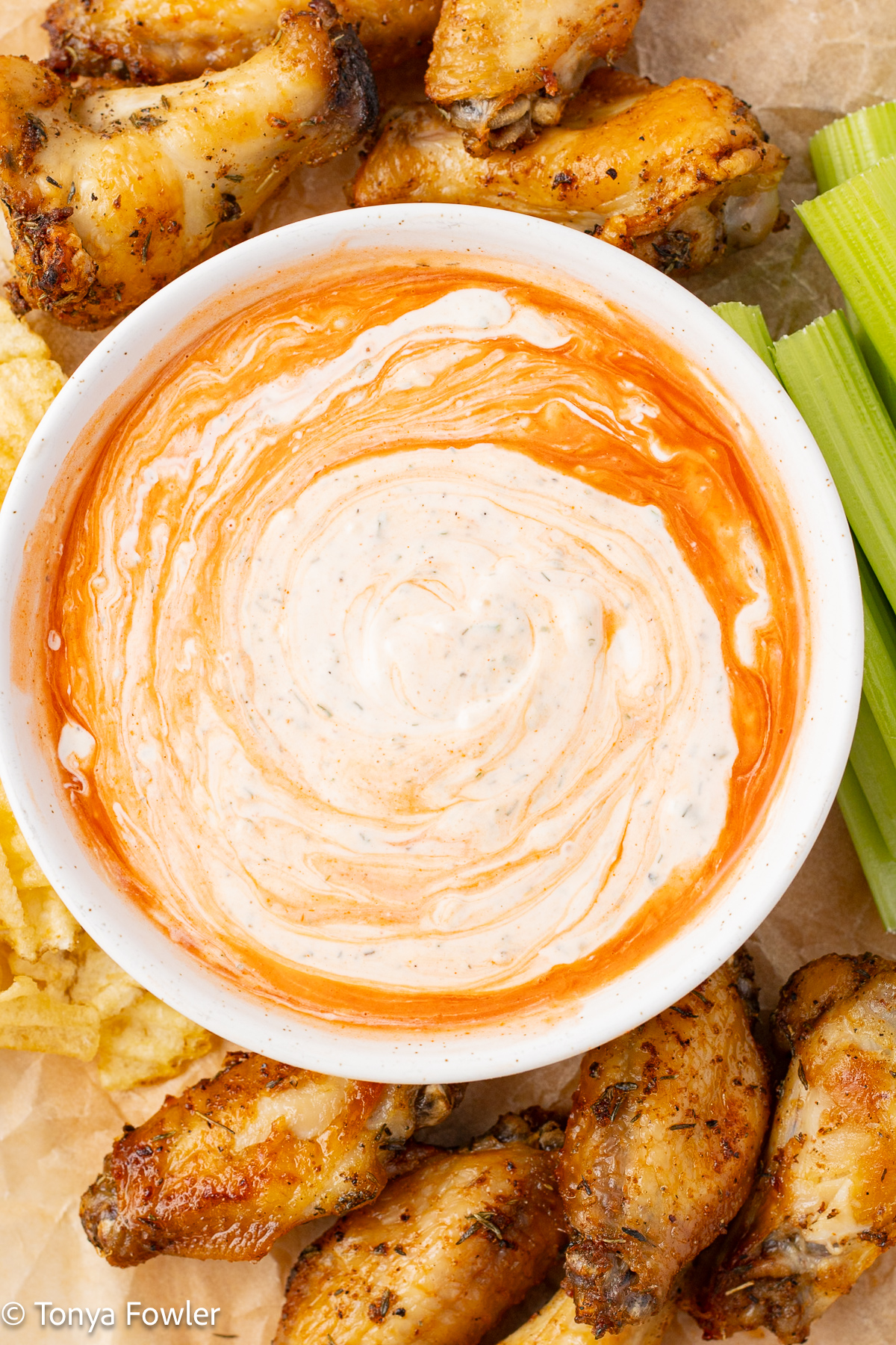 Ranch dressing in a bowl surrounded with chicken wings, potato chips, and celery sticks.
