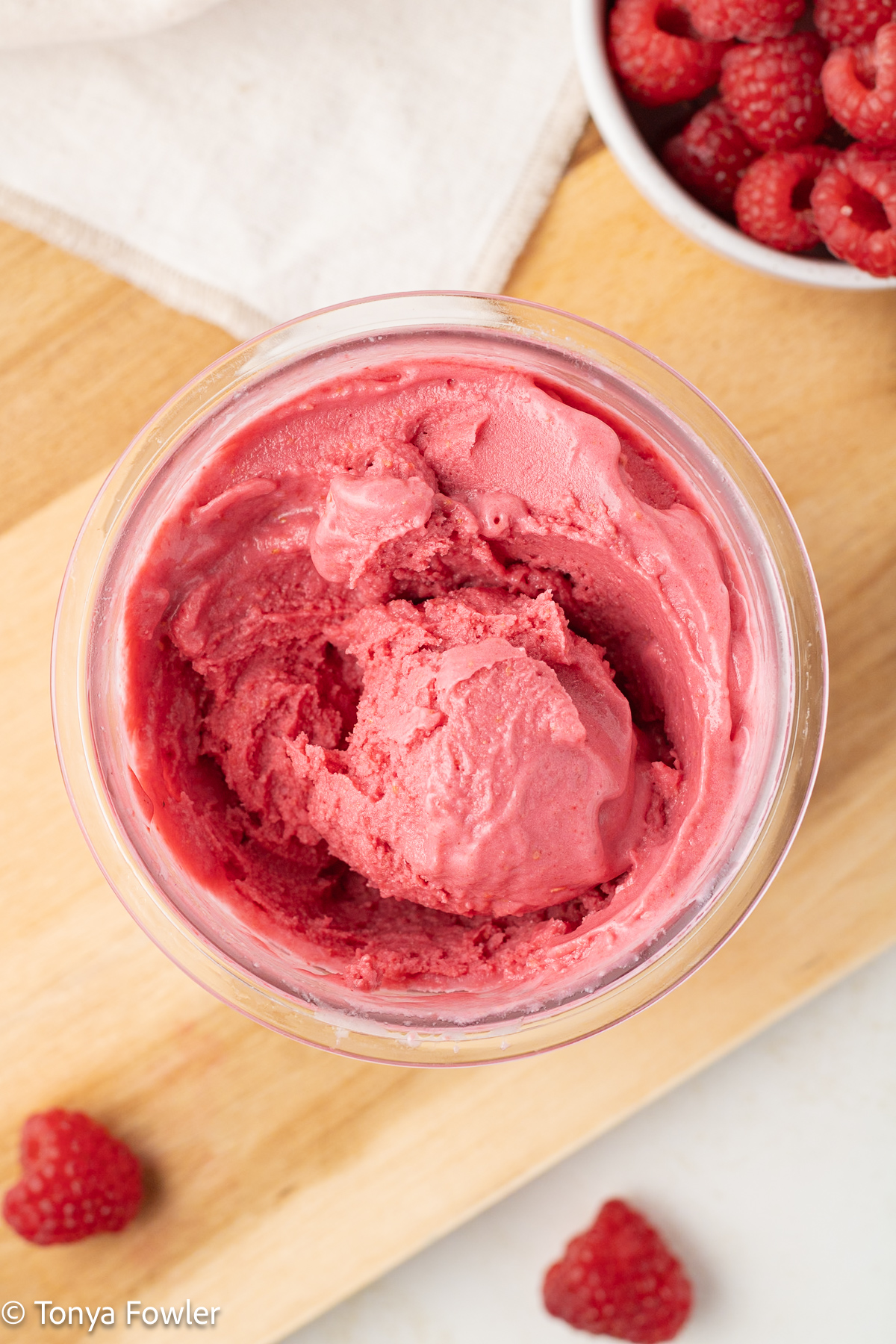 Overhead image of a scoop of raspberry frozen yogurt in a container with fresh berries on the table.