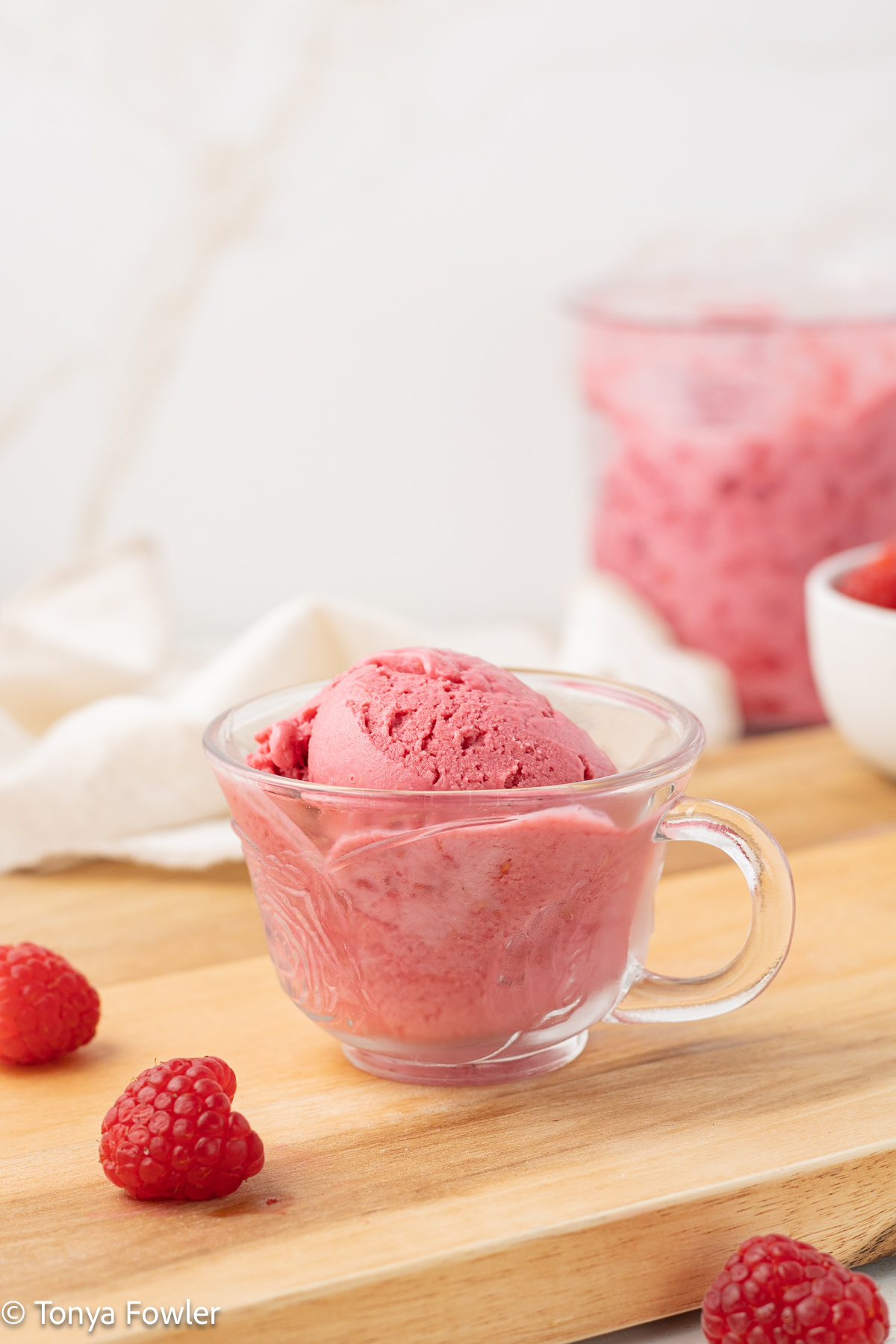 A scoop of frozen yogurt in a cup sitting on a cutting board with fresh raspberries all around.