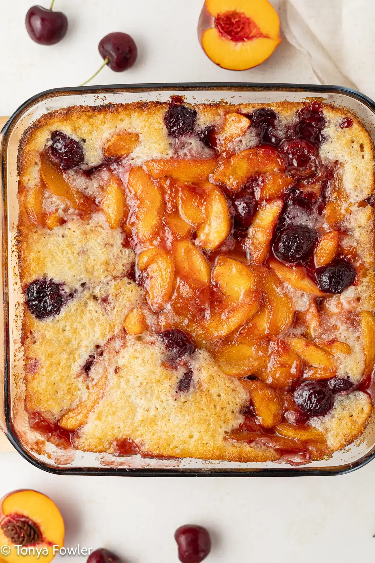 Overhead image of peach cherry cobbler in a baking dish with fresh fruit all around.